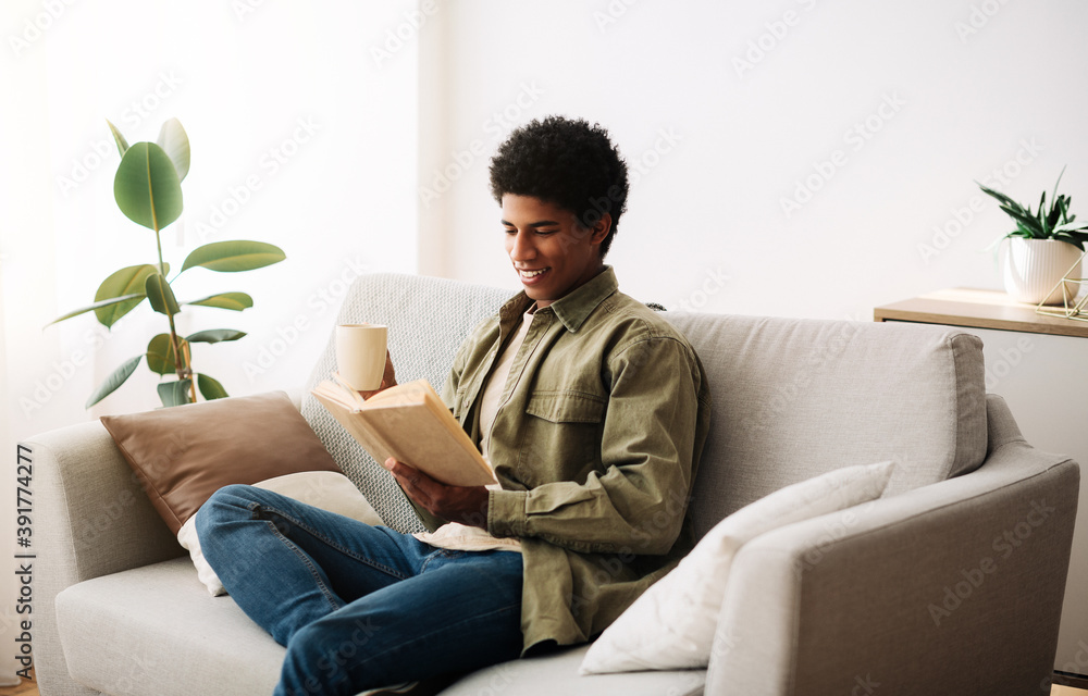 Cheerful African American teenager with cup of hot coffee reading book on sofa at home
