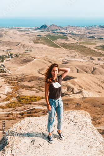 A young beautiful woman with long hair and jeans stands on a rock in the Koktebel valley in Crimea. The landscape of sandy hilly desert. © KseniaJoyg