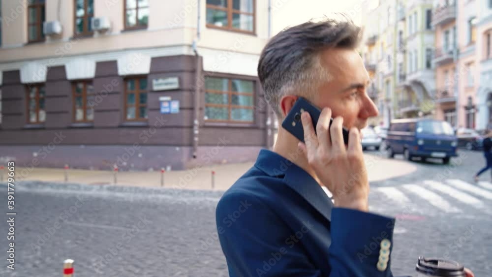 Caucasian handsome middle-aged businessman walking the street, drinking coffee and talking on mobile phone. Man strolling and speaking on cellphone. Sipping hot drink. Busy male calling on telephone. Caucasian handsome middle-aged businessman walking the street, drinking coffee and talking on mobile phone. Man strolling and speaking on cellphone. Sipping hot drink. Busy male calling on telephone.