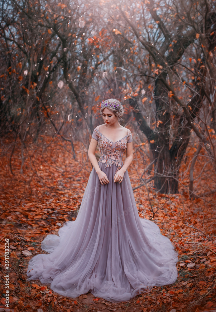 attractive girl princess in long medieval purple dress stands in fairy autumn forest. Silhouette of dark Gothic trees fallen orange foliage. Woman fashion model in image of luxury queen. Crown on head