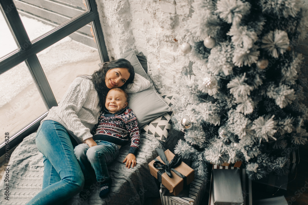 Happy mother and her son lies on windowsill near Christmas tree and gift box.