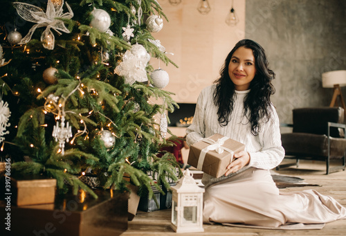 Happy woman sits on the floor and holds gift box near Christmas tree.