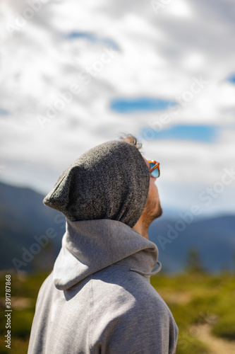 Young white man from behind with sunglasses in beautiful natural setting surrounded by mountains looking up at cloudy sky