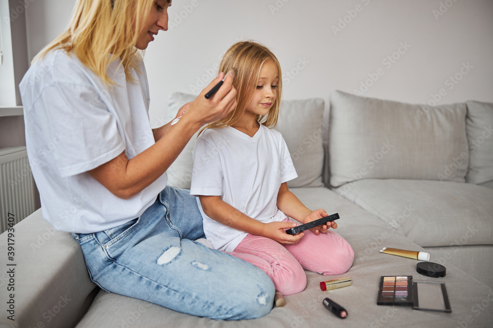 Blonde Caucasian woman in casual clothes having fun with small miss while doing make up Blonde Caucasian woman in casual clothes having fun with small miss while doing make up