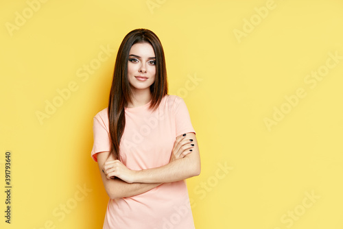 Pretty confident young beautiful woman in casual clothing with crossed arms over yellow background