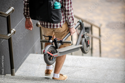 Man Carrying folded E-Scooter in hand on stairs near building. Moving by eco urban transport, modern city concept. Ecological technological lifestyle. Person Carry Electric Scooter In Folded Position