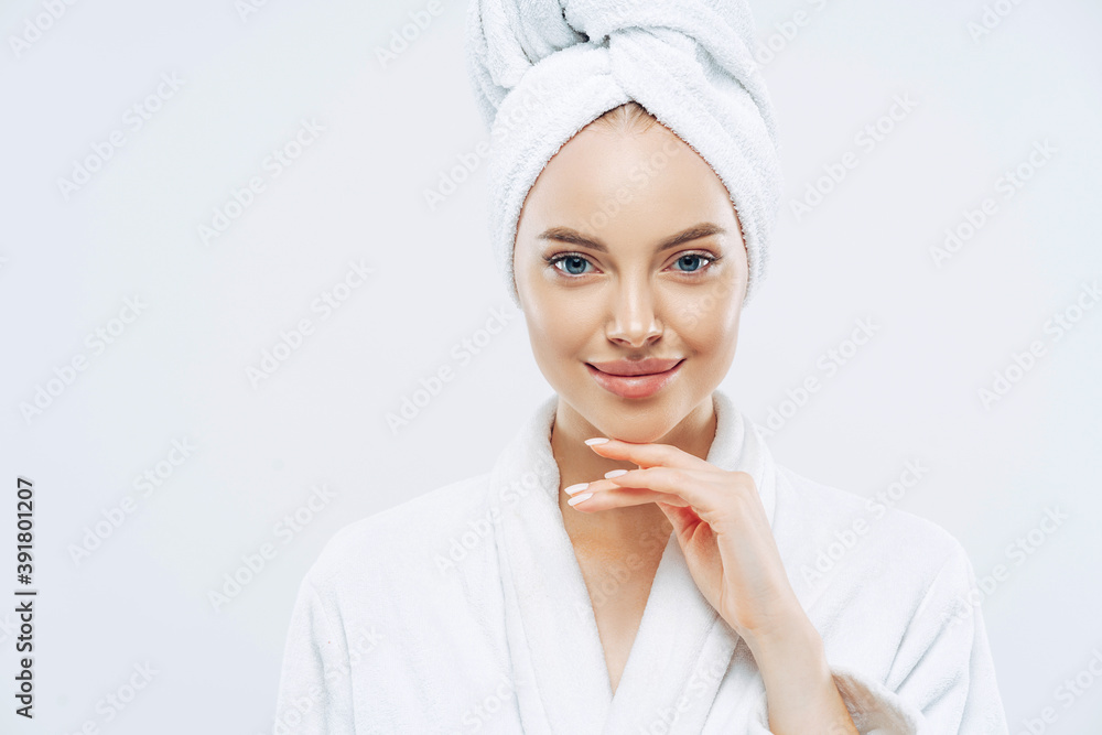 Cropped shot of young good looking European woman looks with calm face, enjoys bath procedures, wears soft white robe, wrapped towel on head, isolated on white background. Morning time, hygiene Cropped shot of young good looking European woman looks with calm face, enjoys bath procedures, wears soft white robe, wrapped towel on head, isolated on white background. Morning time, hygiene