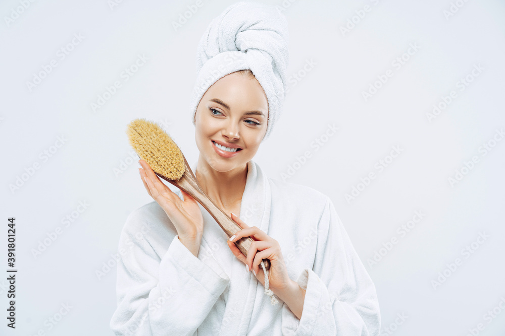Beautiful glad woman with healthy skin and toothy smile holds wooden massage brush for body, wears bath robe and wrapped soft towel on head, stands against white background. Beauty, care concept Beautiful glad woman with healthy skin and toothy smile holds wooden massage brush for body, wears bath robe and wrapped soft towel on head, stands against white background. Beauty, care concept