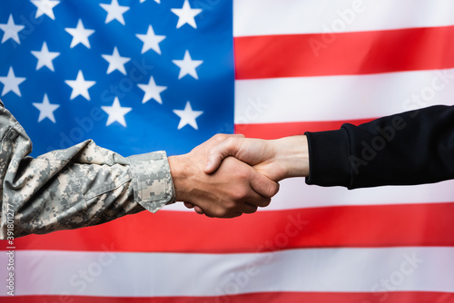 cropped view of soldier shaking hand with civilian man near american flag on blurred background