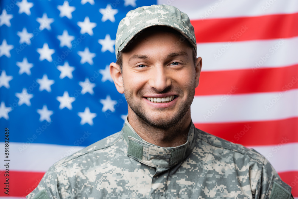 happy military man in uniform and cap smiling near american flag on blurred background happy military man in uniform and cap smiling near american flag on blurred background