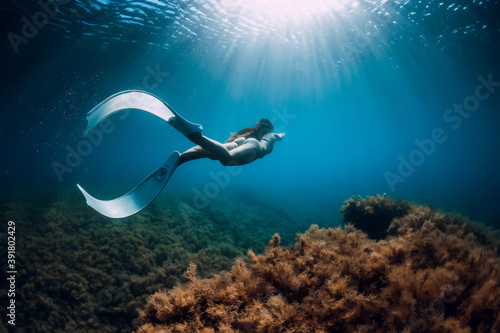 Freediver girl with white fins glides near rock with seaweed and sun rays. Freediving and beautiful light in blue sea