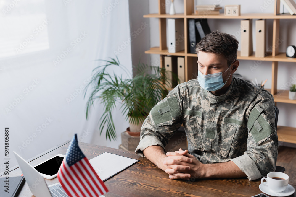 military man in uniform and medical mask sitting with clenched hands at desk with american flag military man in uniform and medical mask sitting with clenched hands at desk with american flag