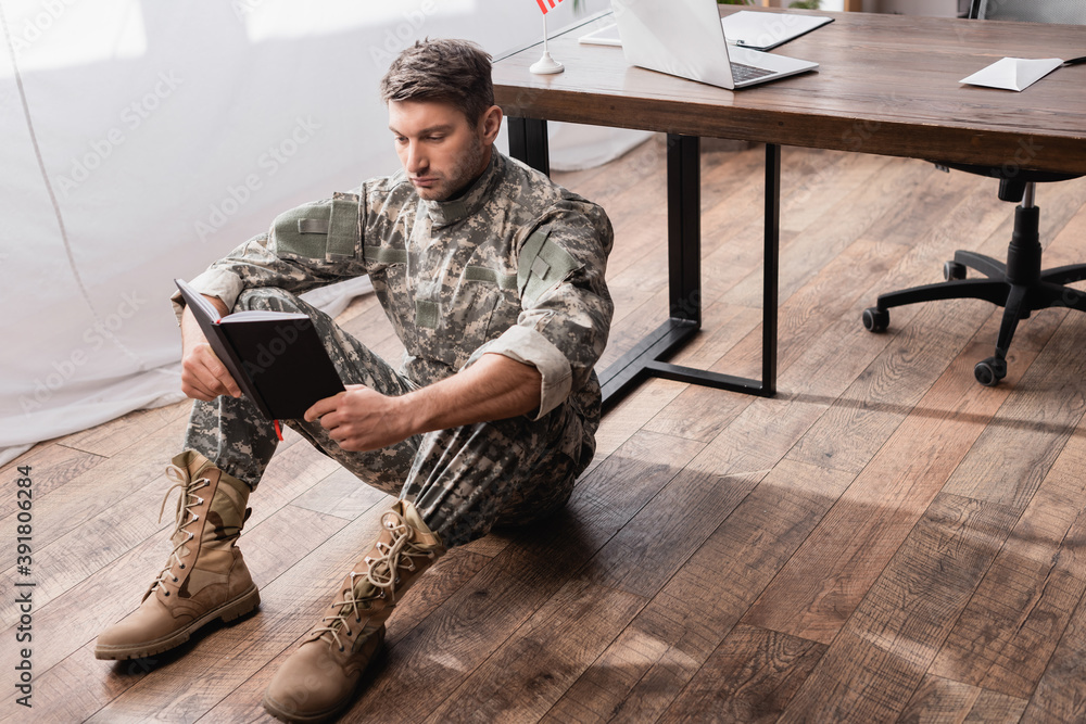 focused military man in uniform holding copy book while sitting on floor near desk