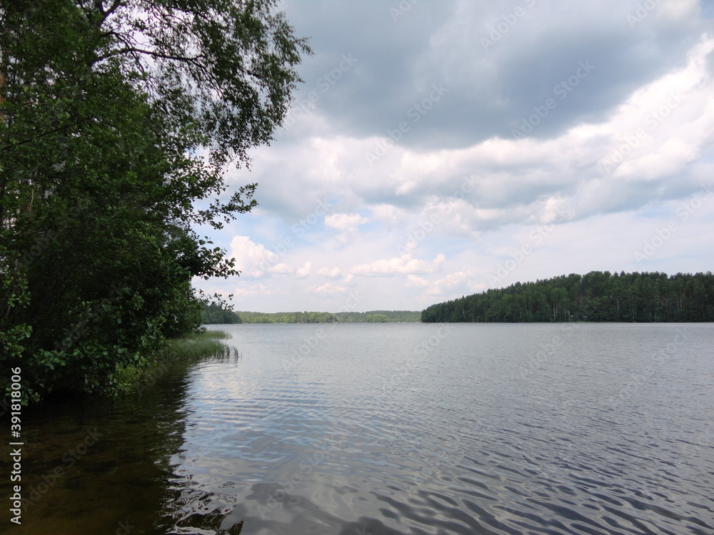 Lake and forest around.
The forest surrounds a beautiful lake with clear water. Trees and bushes grow on the shore. Blue sky with white clouds over the lake and forest. The sky is reflected in the wat