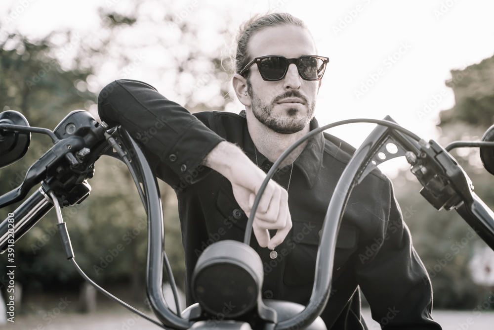Unsaturated photo of a man with sunglasses leaning against the handlebars of a motorcycle outdoors