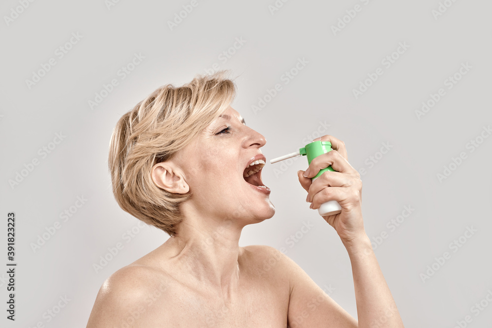 Portrait of attractive middle aged woman using throat spray, standing isolated over grey background Portrait of attractive middle aged woman using throat spray, standing isolated over grey background