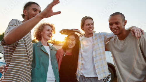 Five casually dressed friends of different nationalities hugging each other having a good time together outside laughing and smiling with their car on a background
