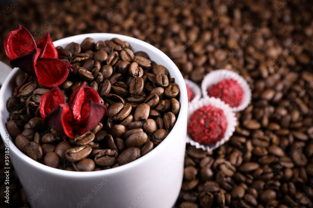 Fototapeta premium White cup full of coffee beans on a coffee beans background with red chocolate candy and red flowers. Morning espresso. Coffee mug.