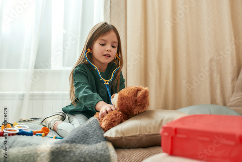 A littile girl taking medical care of her teddybear patient holding a toy stethoscope while sitting on a sofa in a bright room photo
