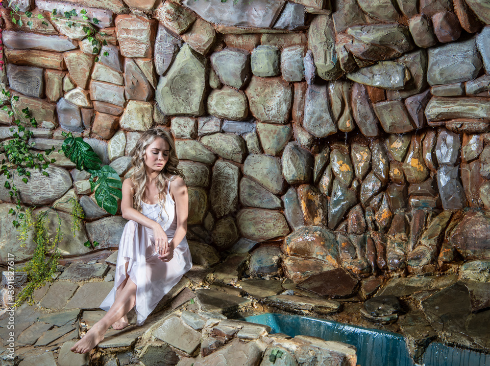 beautiful blonde in a white dress posing near a stone wall with a stream