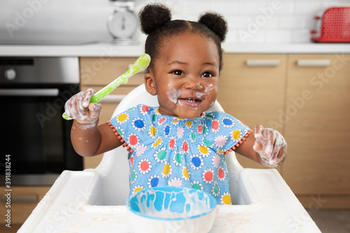 Happy baby girl with messy face eating yogurt with spoon in high chair
