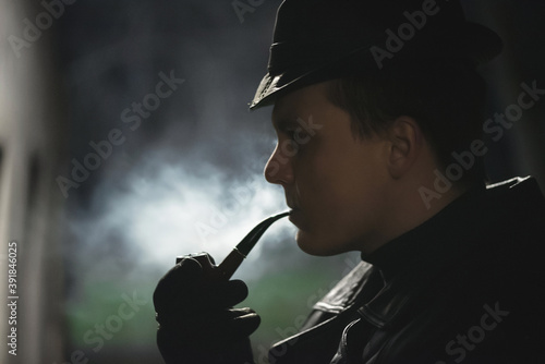 Detective agent in black leather hat and coat smokes a smoking pipe.