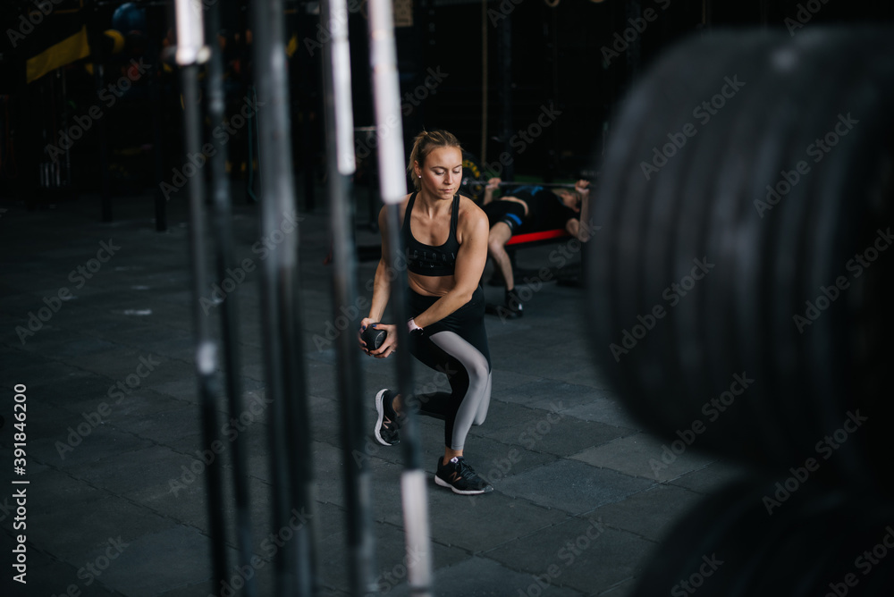 Sporty young woman with perfect athletic body wearing black sportswear working out with kettlebell during sport workout training at modern fitness gym with dark interior. Sporty young woman with perfect athletic body wearing black sportswear working out with kettlebell during sport workout training at modern fitness gym with dark interior.