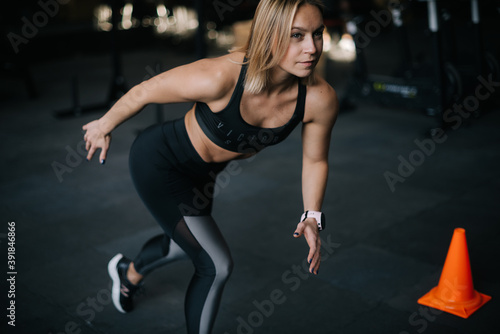 Sporty young woman runner with perfect athletic body wearing black sportswear posing at modern gym with dark interior, dynamic movement.