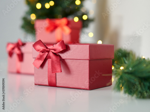 Close up of a red gift box. Beautiful gift box with a red ribbon and a bow. Background with green Christmas tree and lights. Christmas time and gift time © Lukasz