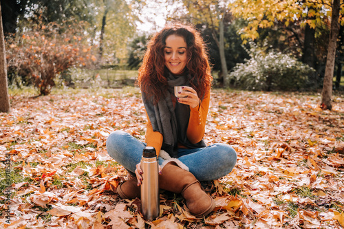 A beautiful brunette woman sitting on the ground surrounded by leaves while enjoying a coffee in the park. She is putting the thermos on the floor. Lifestyle autumnal outdoors