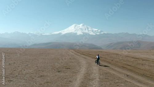 Woman riding on electro e-bike bicycle at mountain plain on snowy peak and valley landscape from drone. Traveler woman bicycling in mountain bike on highlands on snowy Elbrus Dzhily Su landscape. photo