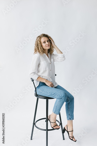 Beautiful blonde girl in white shirt and jeans sitting on a chair on a white background in the studio. © Marina