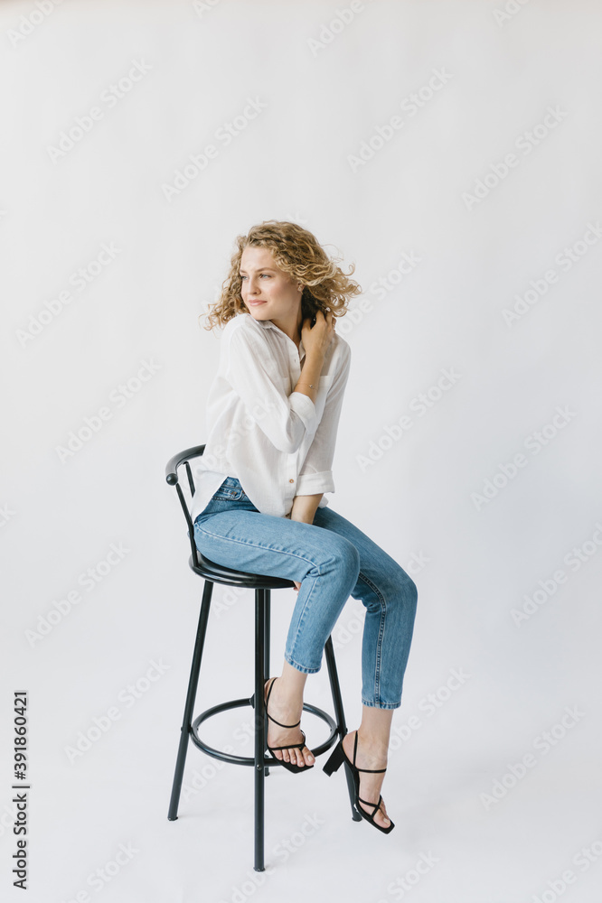 Beautiful blonde girl in white shirt and jeans sitting on a chair on a white background in the studio.