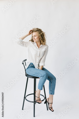 Beautiful blonde girl in white shirt and jeans sitting on a chair on a white background in the studio. © Marina