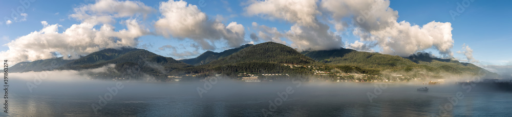 Panoramic shot of mountains covered with clouds and fog in Gastineau Channel, Juneau, Alaska. Golden hour. Blue cloudy sky as a background and boat sailing in the fog in the foreground