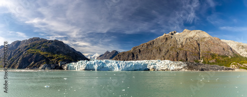 Panoramic view of Margerie glacier surrounded by huge mountains in Glacier Bay National Park and Preserve, Alaska, United States. Snowy mountain peaks and cloudy blue sky in the background