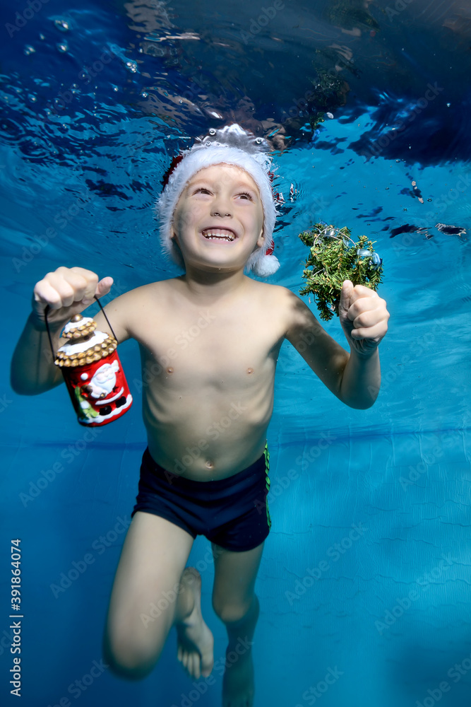 Laughing little boy swims and plays underwater in the pool in a red Santa hat with Christmas gifts in hand on a blue background. Portrait. Vertical orientation of the photo