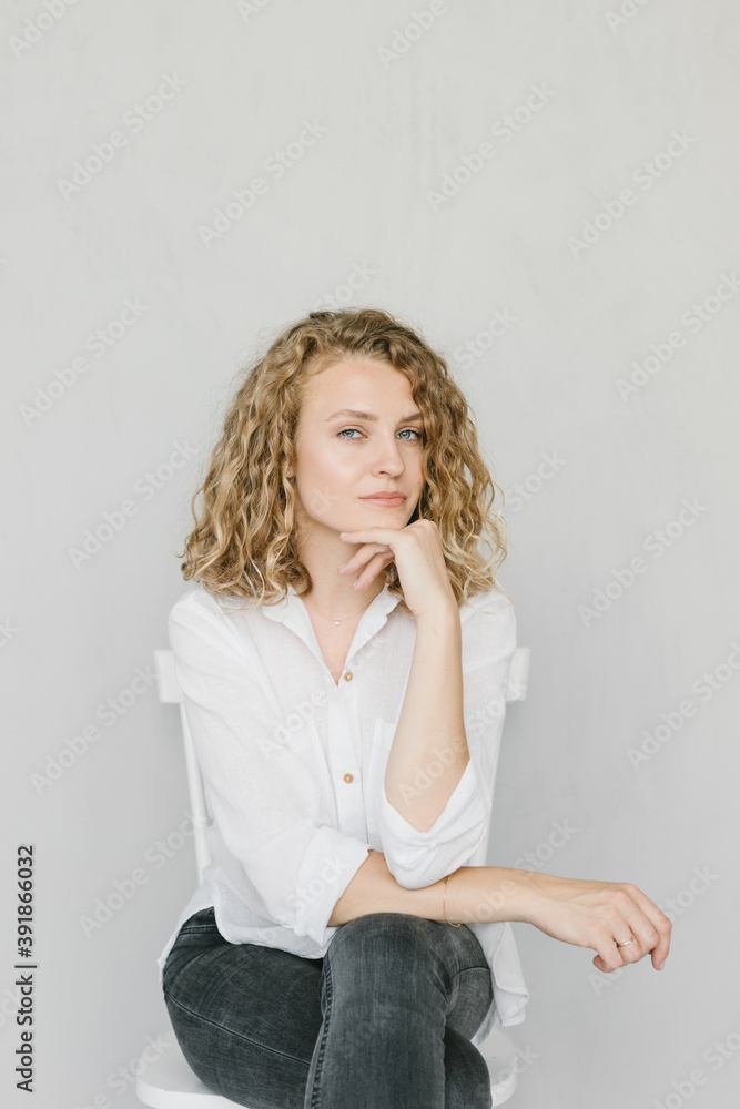 A beautiful blonde girl in a white shirt and jeans is sitting on a chair on a gray background in the studio. A beautiful blonde girl in a white shirt and jeans is sitting on a chair on a gray background in the studio.