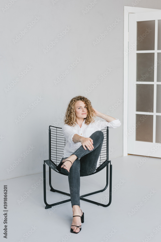 A beautiful blonde girl in a white shirt and jeans is sitting on a chair on a gray background in the studio. A beautiful blonde girl in a white shirt and jeans is sitting on a chair on a gray background in the studio.