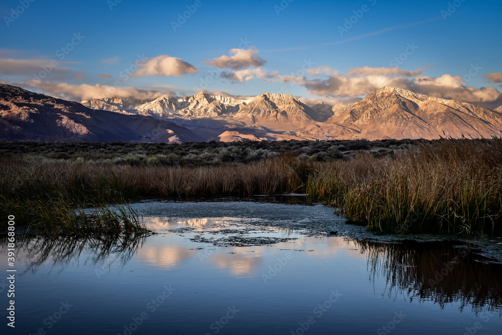 First morning light illuminates mountains with darker valley  wetlands grasses reflection in water