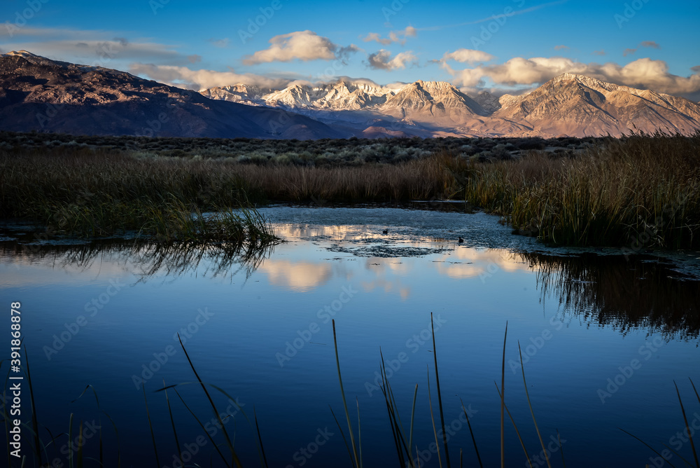 First morning light illuminates mountains with darker valley  wetlands grasses reflection in water