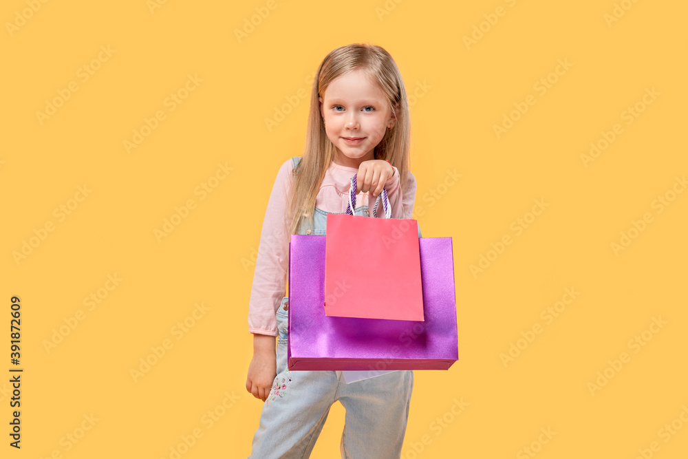 Fototapeta premium A cute girl looks at the camera and holds shopping bags in her hands. Gifts for the holidays. Yellow isolated background.