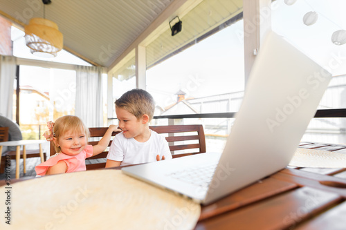 Small smiling kids brother and sister laugh and play laptop, communicate video conference chatting. Spacious cozy home interior.