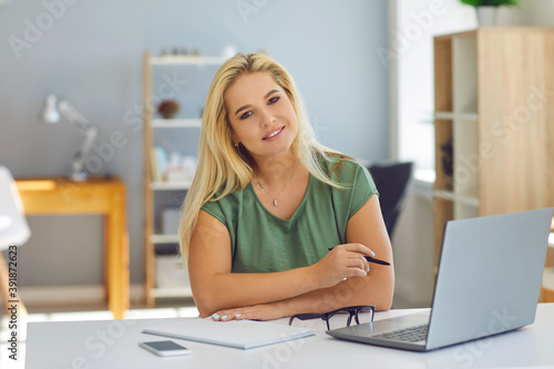 Smiling woman working online with notebook and looking at camera at home or modern office