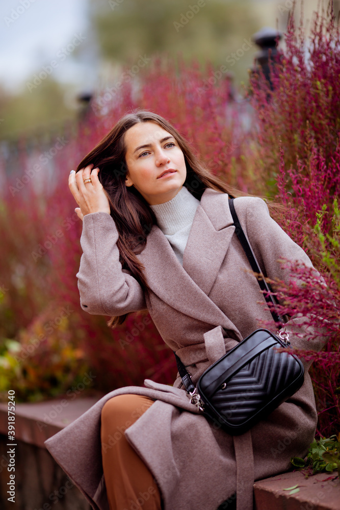 beautiful long-haired brunette in a light beige coat against a background of plants beautiful long-haired brunette in a light beige coat against a background of plants