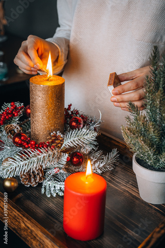 Close up of lighting first candle on Advent Wreath Celebrating Christmas or new year holidays. warm lights, xmas tree winter home interior decorations. hand holding match to fire candle. Vertical