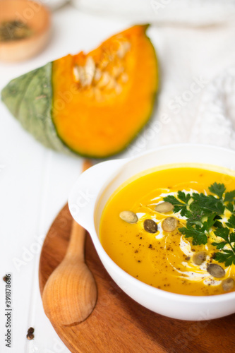 A plate of yellow pumpkin cream soup with cream, herbs and seeds on a white table near raw pumpkin and a wooden spoon.