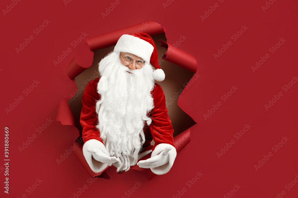 Santa man spread his arms in different directions and look emotionally through a paper hole. Bearded man in santa hat looking through hole on red paper. Santa man spread his arms in different directions and look emotionally through a paper hole. Bearded man in santa hat looking through hole on red paper.
