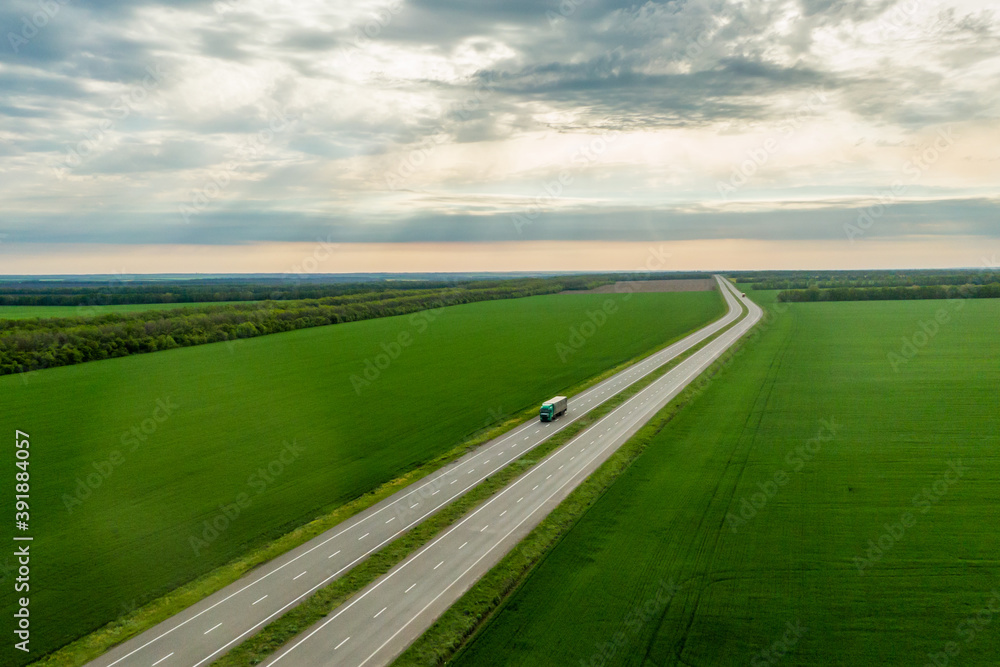 Fototapeta premium green truck driving on asphalt road along the green fields. seen from the air. Aerial view landscape. drone photography. cargo delivery