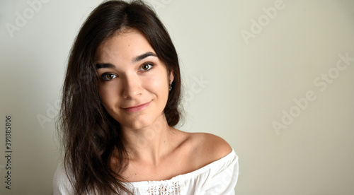 Portrait of young 25 y.o. brunette Russian woman with long hair. In white open shoulder top and looking into the camera on light gray background. Minimum makeup, natural hairstyle. copy space